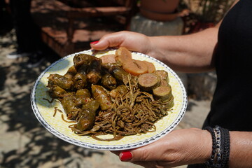 A traditional Cretan plate features vine leaves stuffed with wheat, tender wild greens, zucchini slices, and root vegetables. A woman holds it under the sunlight in an outdoor courtyard.