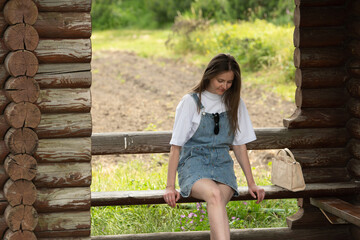 Naklejka premium A young woman sits on a wooden bench in a rustic structure, enjoying a tranquil moment in nature. The surrounding field blooms with greenery during a bright, sunny day