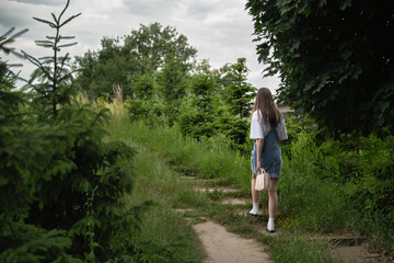 A woman navigates a narrow dirt path surrounded by dense greenery on a cloudy day. She carries a...