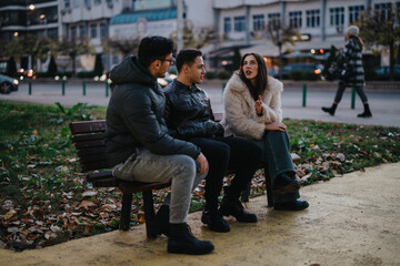 Three friends engage in a friendly discussion while seated on a bench in an urban park. A passerby...