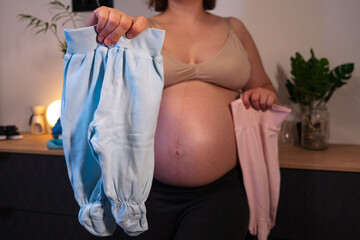 Pregnant woman with big belly in anticipation for the motherhood. Concept of pregnant woman wondering if it a boy or a girl gender with with rompers in hands. Twins