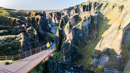 A traveler walks across a metal bridge, gazing at the dramatic cliffs and lush greenery of Icelands...