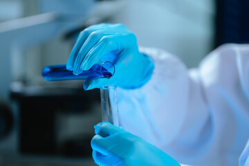 Scientists in lab coats and gloves pour blue liquid from one test tube to another to perform...