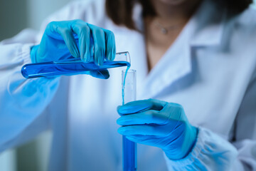 Scientists in lab coats and gloves pour blue liquid from one test tube to another to perform...