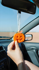 Female hand gripping seasonal pumpkin ornament dangling from car's rearview mirror, creating autumn driving ambiance