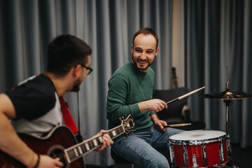 Two adult men creating music in a studio, one playing the drums and the other on guitar, sharing a...