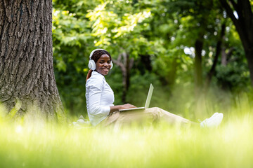 Beautiful African businesswoman sitting on grass and working on laptop computer in city park.