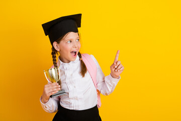 Excited schoolgirl celebrating achievement with trophy wearing academic cap on bright yellow...