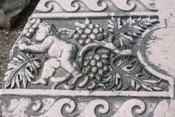 Bas-relief on the surface of a stone block in the ancient city of Perge in Turkey. An image of an angel and grapes on a stone surface. Grape leaves and berries next to an angel on a stone.
