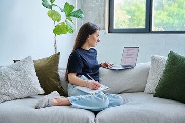 Young student girl study online, sitting at home on couch with laptop, notebook