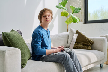 Handsome serious teenage boy sitting at home on the couch typing on laptop