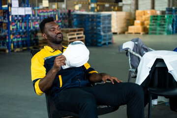 African male warehouse worker sitting back relaxed while holding helmet taking rest after physical...