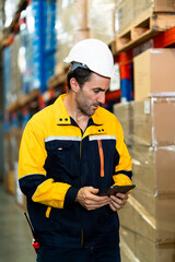 White adult male warehouse worker smiling while checking tablet and walking along packed boxes and...