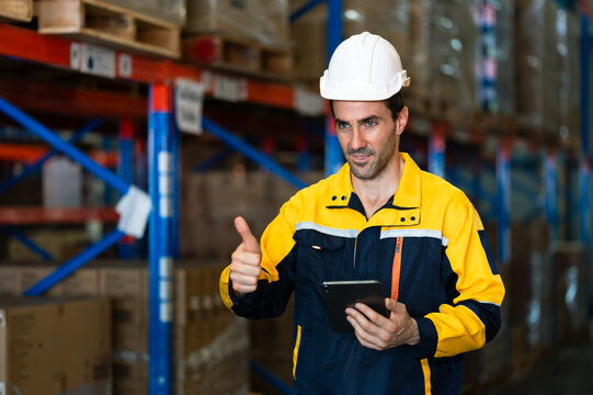 Caucasian male warehouse supervisor holding tablet smiling with thumbs up during successful stock check operation inside logistic facility filled with organized packages and pallet shelves