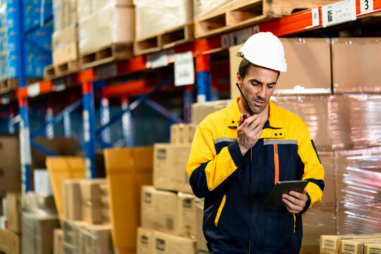 White adult male worker in logistics warehouse analyzing data on tablet while touching chin, dressed in uniform and helmet surrounded by packed boxes and tall racks, inside logistics warehouse.