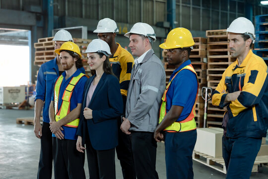 Multiracial warehouse staff standing in line listening attentively to briefing inside industrial storage zone wearing diverse uniforms and helmets showing multicultural workforce unity