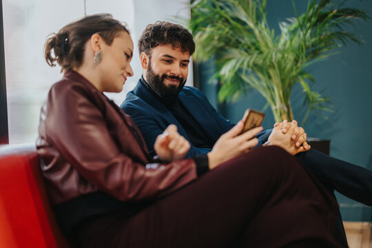A man and a woman conversing while looking at a smart phone. Features informal business attire, warm expressions, and a comfortable lounge environment with plants enhancing the ambiance.