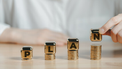 Gold cubes with letters PLAN on stacked coins and hand arranging blocks symbolizing financial...
