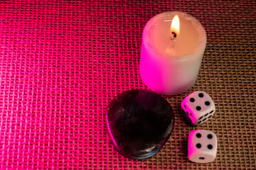 A lit candle, dice, and labradorite rest on burlap, bathed in soft pink light. A mystical flat lay...