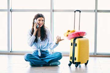 Charming woman sitting at the airport holding a phone next to a yellow suitcase, ready for her next...