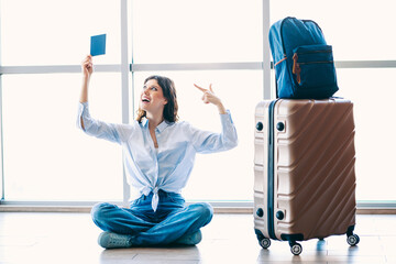 Young woman sitting at airport holding a ticket near luggage ready for travel adventure enjoying...