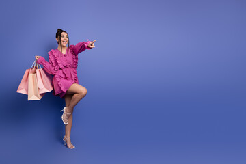 Smiling woman wearing stylish ruffle dress holding shopping bags on a purple background with joyful...