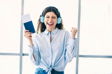 Young female traveler excited with tickets wearing headphones in bright airport terminal during...