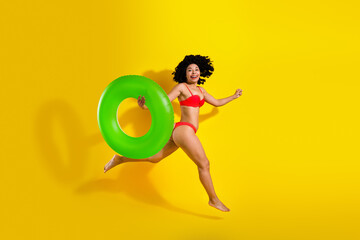 Positive woman in a red bikini holding a green pool float on yellow backdrop, representing summer,...