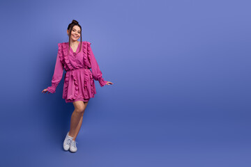Charming young woman in a pink ruffled dress posing against a vivid purple backdrop showcasing...