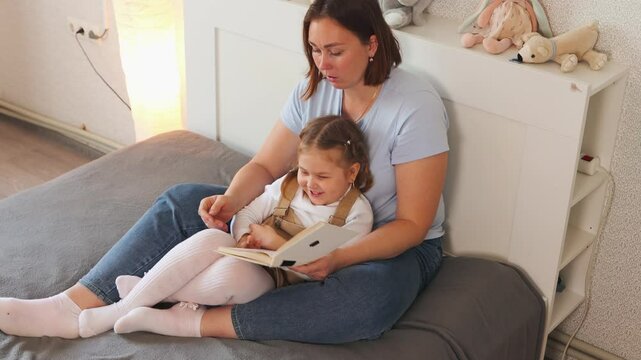 Beautiful woman and her little girl enjoy reading a story together in bed. Concept of child learning, maternal care, and speech development at home. Top view