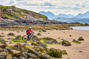 cheerful senior woman riding her electric mountain bike at a rocky beach of the scottiish Atlantic...