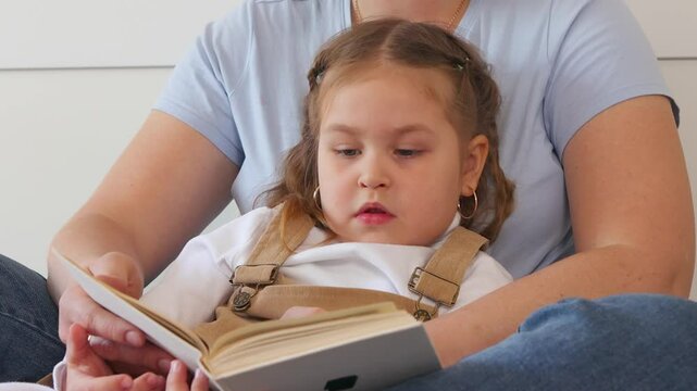 Young Caucasian mother reads a book to her daughter while cuddling in bed. Family bonding, early education, and emotional development in a cozy bedroom environment. Close up.