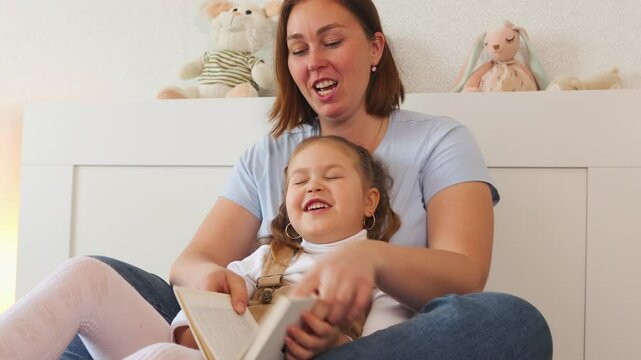 Young Caucasian woman hugs her little daughter while reading a book together in a cozy bedroom. Concept of early education and child development at home