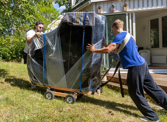 Two movers transporting a grand piano on a four-wheeled dolly across a garden toward a house on a...