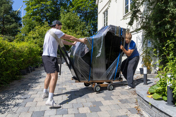 Two movers handling a wrapped grand piano on a dolly, navigating a paved path outside a residential...