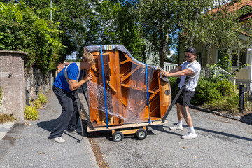 Two movers transporting a grand piano on a small road, using a dolly and lifting straps. The instrument is wrapped in plastic and blankets for protection. © Christo