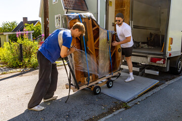Two movers loading a partially wrapped grand piano onto a truck using a hydraulic lift on a...