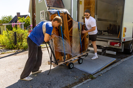 Two movers loading a partially wrapped grand piano onto a truck using a hydraulic lift on a residential street. - Powered by Adobe