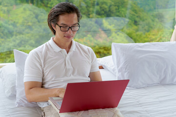 Young asian man with glasses working remotely on a laptop while sitting on a cozy bed with scenic mountain views through large glass windows. Modern work-from-anywhere lifestyle in green season.