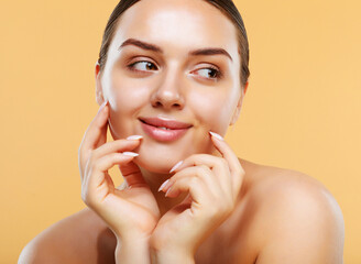 Beautiful young woman with clean fresh skin touching her face over beige background.