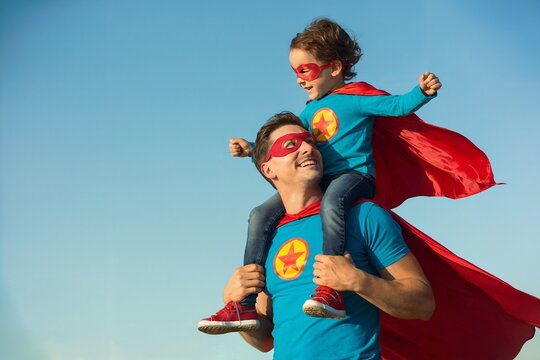 Father And Son In Matching Superhero Costumes Playing Outdoors Under Bright Sky
