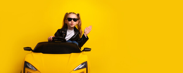 Stylish young girl wearing sunglasses and leather jacket drives a yellow toy sports car against vibrant yellow background.