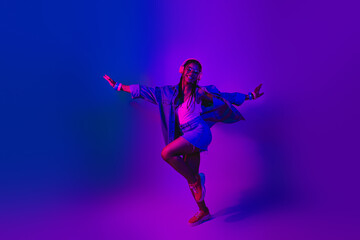 Stylish young woman enjoying a dance in vibrant neon lighting wearing denim jacket and headphones,...