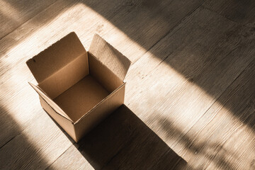 Open empty cardboard box on wooden floor with sunlight casting dramatic shadow, minimalist concept for delivery, shipping, moving, or storage, evoking anticipation and simplicity
