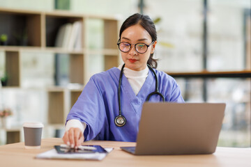 Female medical intern in blue medical scrubs e-learning on laptop computer at the ordination,...