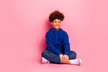 Smiling young boy in a blue hoodie sitting against a pink background showcasing casual fashion and...