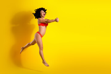 Positive woman in red swimsuit jumping joyfully against a vibrant yellow background expressing...