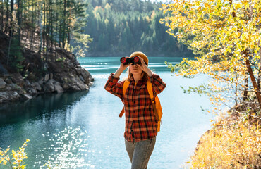 Woman traveler wearing orange backpack and plaid shirt is observing nature with binoculars by serene lake surrounded by trees, essence of outdoor exploration and adventure, birdwatching