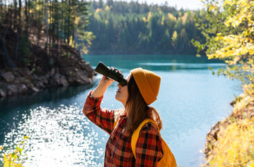 Young woman hiker ecologist in orange beanie and plaid shirt using binoculars by serene lake,...