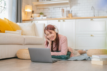 Smiling young freelancer woman wearing headphones, lying on the floor near the window with a...
